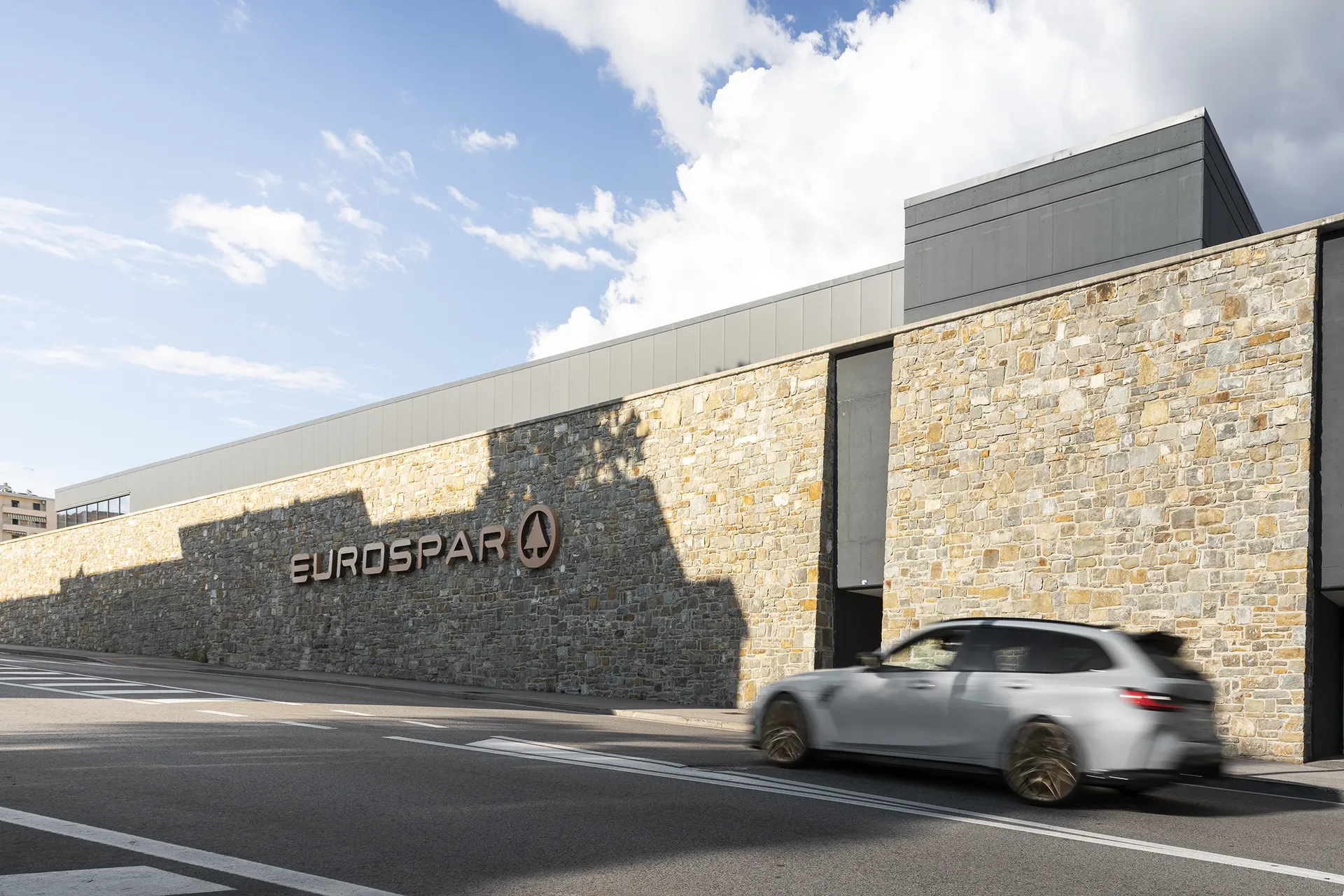 Facciata in pietra di un edificio con insegna "EUROSPAR" e un’auto grigia in movimento su strada asfaltata.
