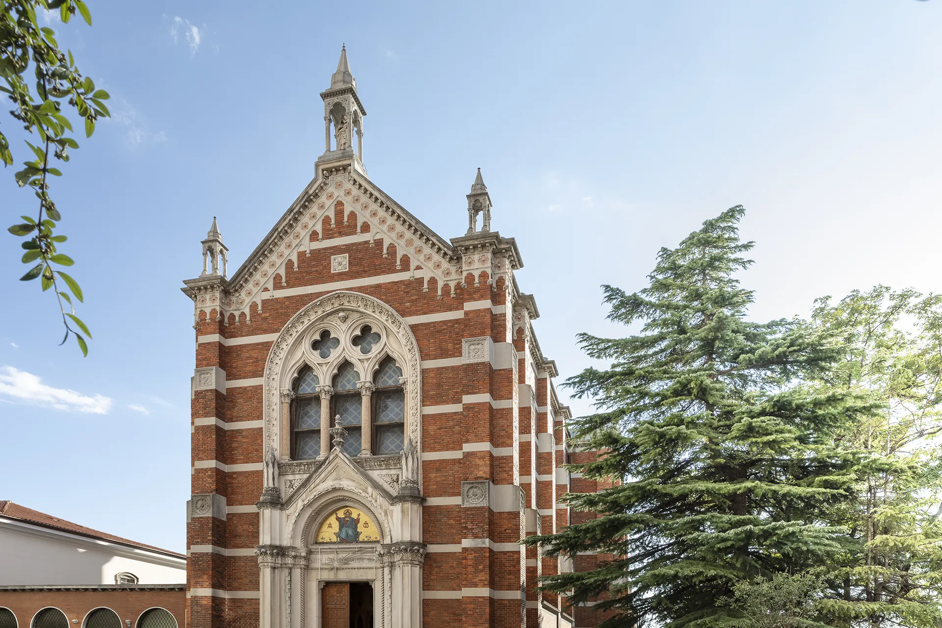 Facciata di chiesa in mattoni rossi e pietra chiara, con portale decorato e vetrate gotiche, affiancata da alberi verdi.