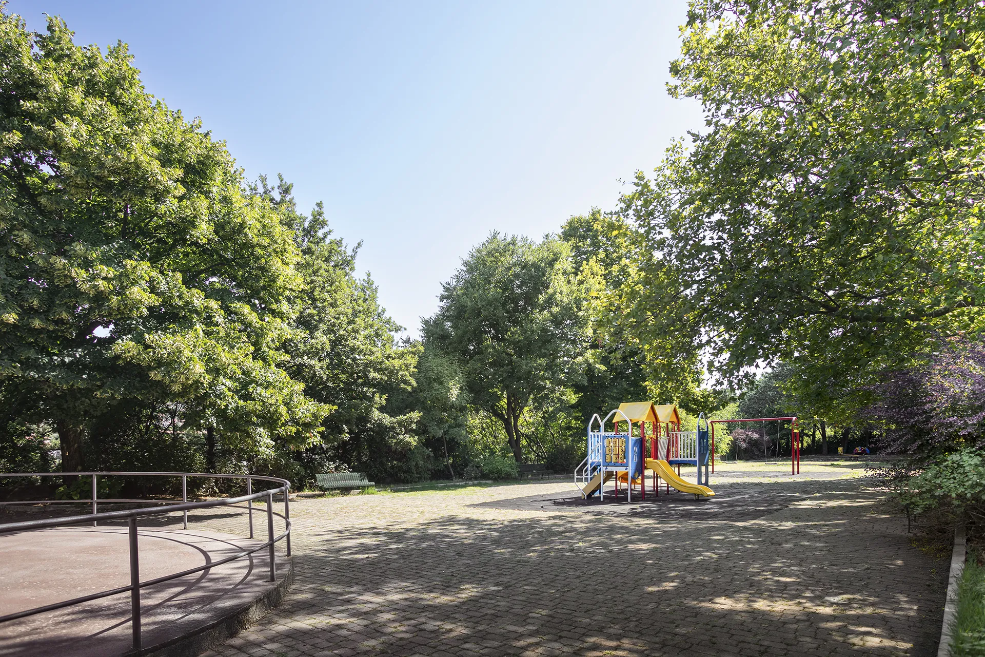 Area giochi per bambini in un parco alberato a Trieste, con scivolo colorato e altalene.